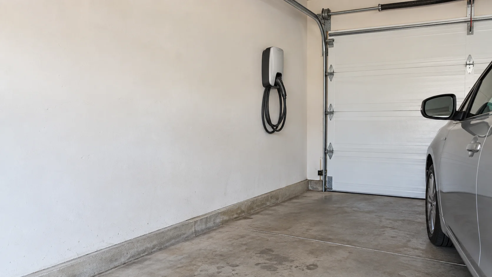 Illustrative everyday home garage with painted drywall, a concrete floor, sectional garage door, wall-mounted EV charger and parked silver car